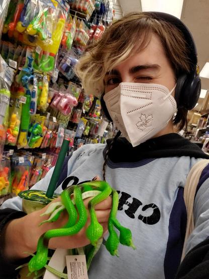 A rare out-of-drag photo of the artist usually known as Austin Tacious. She's in a dollar store holding up a handful of bright green rubber snakes, with a beautifully colour-coordinated matcha bubble tea under one arm. She's winking, wearing headphones and a white mask, and has brown hair that's a little bleached at the ends.