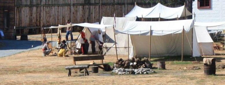 Re-enactors example of an HBC fur trade camp, with canvas tents for the gentlemen and cook fires for all. To the left men talk under a canvas fly. In the background the palisades of replica Fort Langey.