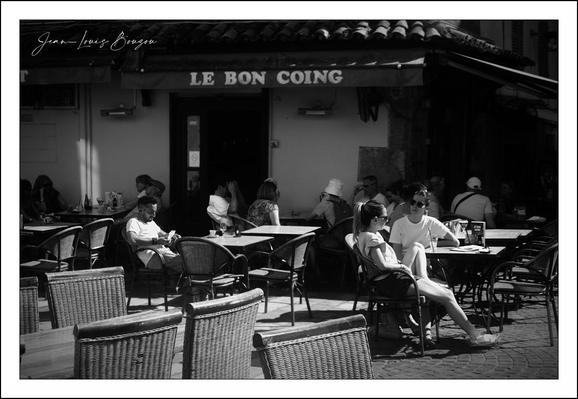 The image captures a lively café scene outside "Le Bon Coing" depicted in soft black and white tones. The café has a charming, rustic vibe, suggested by its tiled roof and warm, inviting facade.
Gathered around the tables are patrons enjoying their time—some engaged in conversation while others are lost in thought. The arrangement of the café chairs is casual and inviting, with a few tables prominently featuring cups and plates, hinting at leisurely meals and drinks.
The play of light and shadow creates a dynamic atmosphere, enhancing the social interactions among the visitors. There's a relaxed yet vibrant energy that suggests enjoyment and community. The black and white aesthetic adds a touch of nostalgia, evoking the classic European café culture where time seems to slow down, inviting moments of connection and reflection.
This image beautifully encapsulates moments of everyday life, where each individual contributes to the collective ambiance of the space, creating a sense of belonging and warmth.
