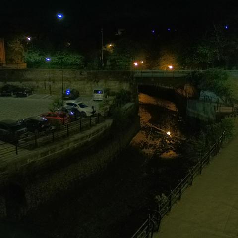 Photograph of the tidal stretch of Ouseburn River at south side of Ouseburn Bridge in Newcastle upon Tyne. To the left (south-west) of the bridge is Cut Bank, and to the right (north-east) is Byker Bank. In the foreground is the pedestrian Riverside Walk and the photo also spans some surrounding built-up areas and trees, with sky in the background. Still image captured from webcam at 03:12 hrs on Friday 2 May 2025.