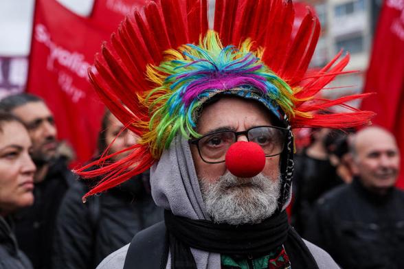 A person wears a clown nose and a red feather headdress as people march to celebrate May Day.