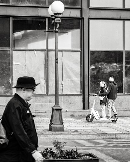 Black and white photo of a man with hat in the foreground and two people riding a scooter in the background