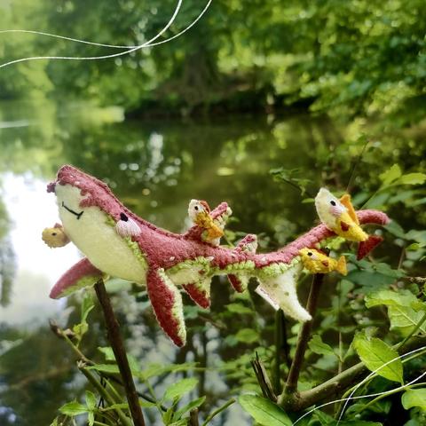 A nurse shark felt plushie photographed in front of a lake. It’s back is a dark vintage pink and it’s belly is ivory, both colors are separated by a light green stripe. Around it, small fishes are keeping it company on its swim.