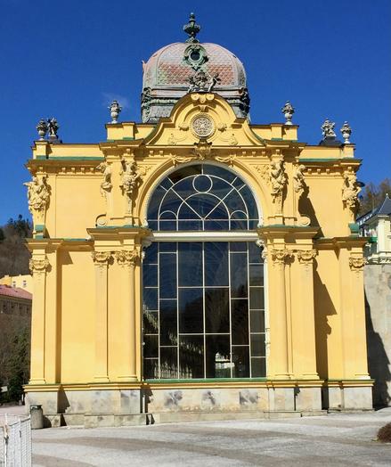 Ein prächtiger Kolonadenbau im Kurbad von Karlovy Vary. Das Gebäude ist im gelben Farbton gehalten und ein großes Fenster ist in der Front.

A magnificent colonnade building in the spa town of Karlovy Vary. The building is yellow in colour with a large window in the front.
