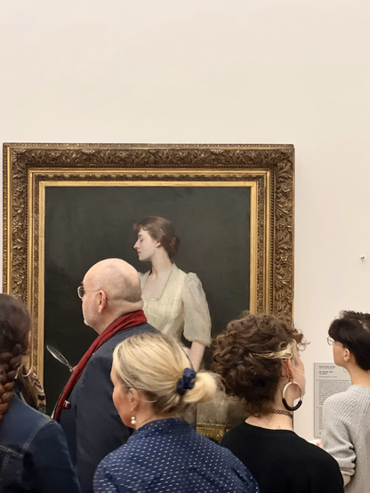 Das Gemälde einer brünetten Frau im weißem Kleid hängt an einer weißen Wand. Das Gemälde hat einen auffälligen Goldrahmen. Vor dem Gemälde befinden sich mehrere Ausstellungsbesuchende. 