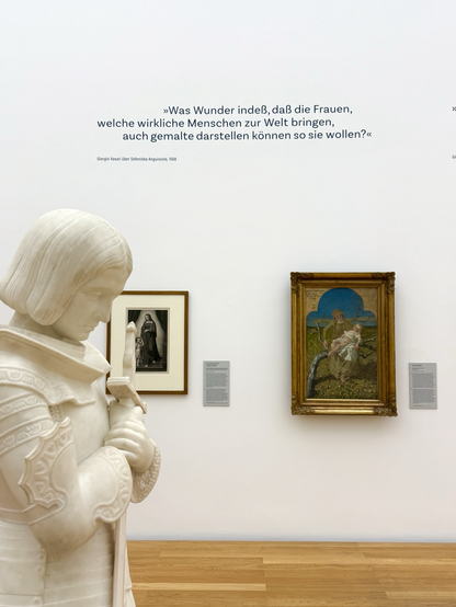 Eine weiße Mamorskulptur steht links in einem Ausstellungsraum. Im Hintergrund hängen zwei Gemälde an einer weißen Wand. Das linke Gemälde ist schwarz-weiß, das rechte gold-blau. Auf beiden sind eine Madonna und ein Jesuskind dargestellt. An der Wand ist ein Zitat angebracht: „Was Wunder indeß, daß die Frauen, welche wirkliche Menschen zur Welt bringen, auch gemalte darstellen können so sie wollen?“ 