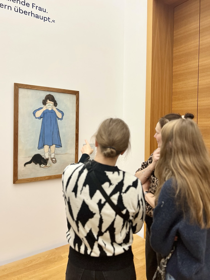 An einer Wand hängt ein Gemälde eines trinkenden Mädchens. Zu ihren Füßen sitzt eine schwarze Katze, die aus einer Schale trinkt. Das Mädchen trägt ein blaues Kleid. In ihrer Hand hält sie eine weiße Tasse, aus dem sie trinkt. Vor dem Gemälde stehen zwei weiblich gelesene Personen. Sie blicken auf das Gemälde.