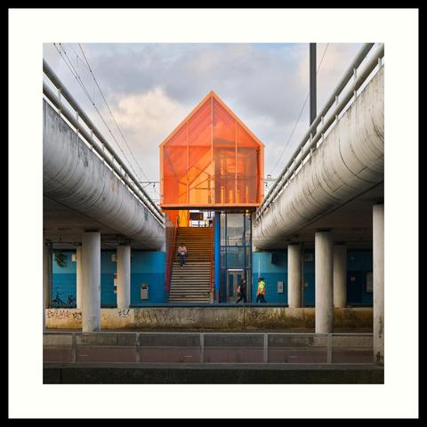 colour - This square format image shows the lowered main entrance to Moerwijk train station. A two-toned blue wall enters from both sides with a staircase and lift in the middle, protected from the elements by an orange-coloured and transparent roof structure. Concrete supports for the tracks come in from both sides. Photographed when the sunlight briefly broke through on this rather overcast morning.