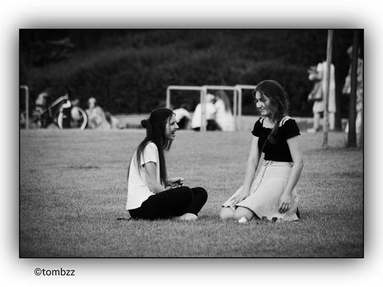 In the black-and-white photograph, two girls are sitting on the grass in the foreground. One has long, loose hair and is wearing a light-colored shirt with dark pants. The other has her hair tied in a braid and is dressed in a dark blouse and a light skirt. Both seem to be engaged in conversation or observing their surroundings. In the background, a group of people is visible—some sitting on benches, others standing or chatting. Several bicycles are leaned against park benches, suggesting that the place is popular among cyclists. The trees and greenery in the distance create a peaceful atmosphere of an urban park or recreational area.