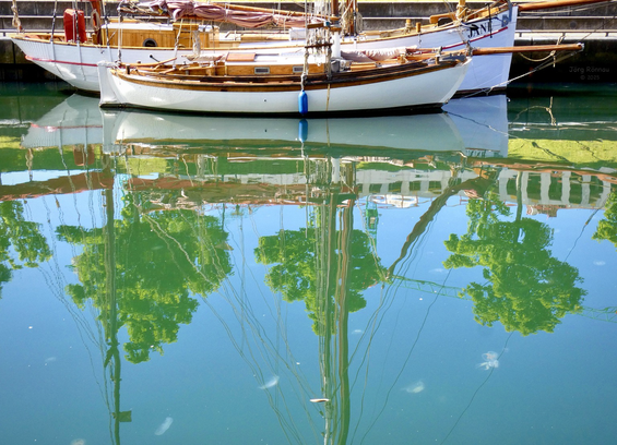 Das Bild zeigt zwei hölzerne Segelboote, die in einem ruhigen Hafenbecken liegen. Ein kleineres Boot liegt vor einem größeren. Die Boote spiegeln sich klar im Wasser, das die grünen Kronen der Bäume an den Ufern zeigt, sowie den hellen Himmel. Der Spiegelungseffekt erzeugt eine fast symmetrische Komposition. Das Bild strahlt Ruhe und Stille aus.
