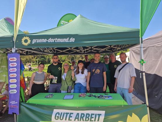 Zehn lächelnde Menschen stehen unter einem grünen Pavillon der Dortmunder Grünen im sonnigen Westfalenpark. Der zweite von links ist Benno Beckmann. Vor ihnen steht liegt ein Infotisch mit Broschüren und Buttons. Ein Banner mit „GUTE ARBEIT“ ist sichtbar. Der Pavillon spendet willkommenen Schatten – die Stimmung ist offen, solidarisch und engagiert.