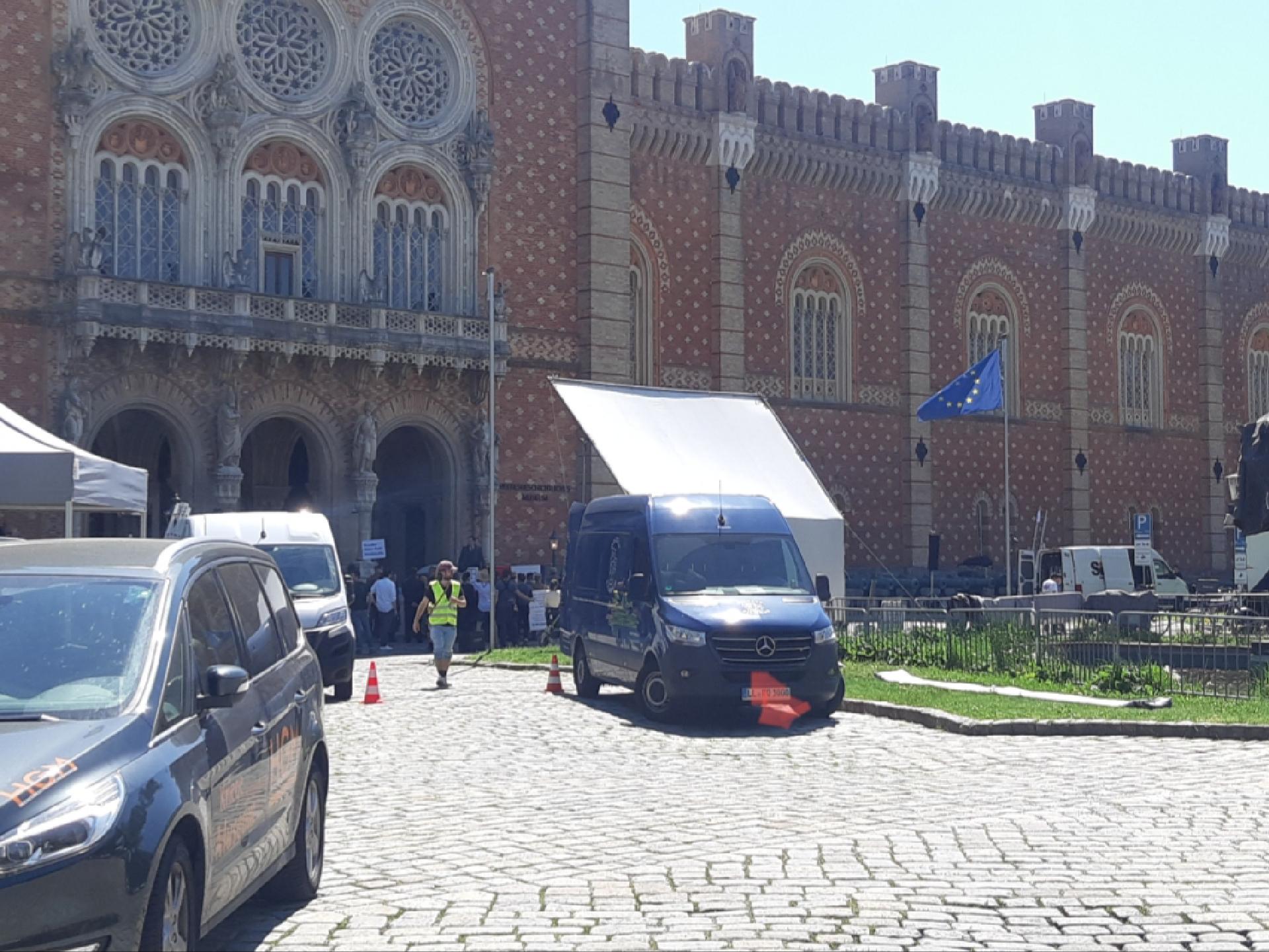 Vorplatz, darauf parkende Trucks, im Hintergrund ein Gebäude mit Menschen davor. 1 Person steht hinter einem Pult neben dem Eingang und spricht zur Menge, die Tafeln oder Transparente mitführt. Davor Kameras