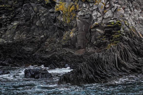 Rocky seaside. A detail of grey rocks, dark grey where they are wet, with some green and orange moss. The water is blue and swirly with some white foamy heads (what is it called in English?). A lot of structure through the photo. The rocks are former lava flows. 