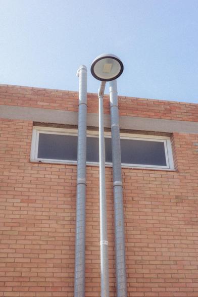Photograph of two pipes and a lamp peeking out from the wall of an industrial building against a blue sky. The wall is made of brick and has an elongated window, partially covered by the pipes and the lamp.