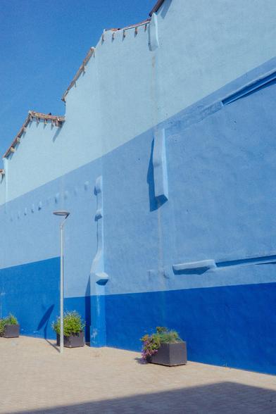 Photograph of a facade painted in three different shades of blue against a very blue sky. There is also a lamp and three planters on the floor.