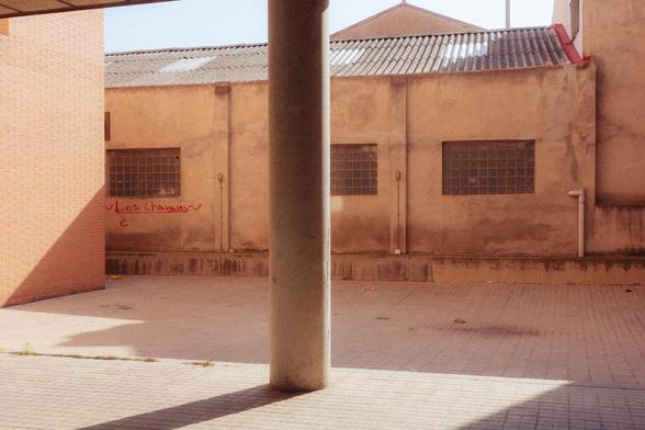 Photograph of a covered courtyard with a column. In the background, a poorly maintained industrial building with graffiti reading "Los chavales" (The Kids) can be seen. The light, shadows, and roofs have very angular shapes, contrasting with the circular column.