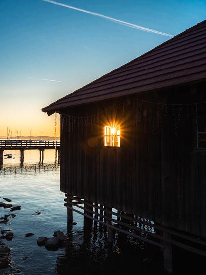 This image shows a peaceful lakeside scene at sunset. In the foreground is a wooden boathouse or cabin built on stilts over water. The structure has dark wooden walls and a brown shingled roof. A window on the side of the building is capturing the setting sun, creating a dramatic starburst effect as the sunlight shines directly through the glass.

The boathouse stands in calm water where small rocks are visible beneath the surface. To the left, you can see part of a wooden pier or dock extending into the water. In the background, there are what appear to be sailboat masts visible near the shoreline.

The sky is a beautiful gradient, transitioning from deep blue at the top to golden yellow near the horizon where the sun is setting. A single contrail from an airplane cuts across the blue sky. The overall atmosphere is serene and golden, capturing that magical moment when day transitions to evening over a tranquil body of water.