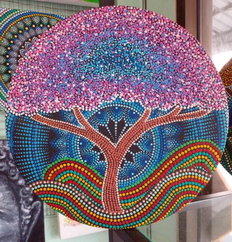 a psychedelic scene, done in small dos of paint, it is a pink and purple and blue -leafed tee with a large blue mandala behind it, a multi-colored wavy ground undernearh it 