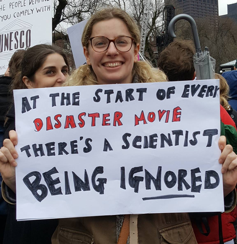 Woman holding a protest sign: At The Start Of Every Disaster Movie There's A Scientist Being IGNORED