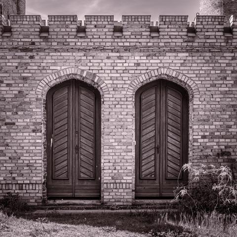 Two wooden doors in a crenellated brick wall, with the foreground overgrown in weeds. Steam Engine House, Babelsberg Palace, Potsdam, 1 May 2025. Nikon D5600, Nikkor DX 35 mm ƒ1.8G, ISO 100, ƒ5, 1/320s
