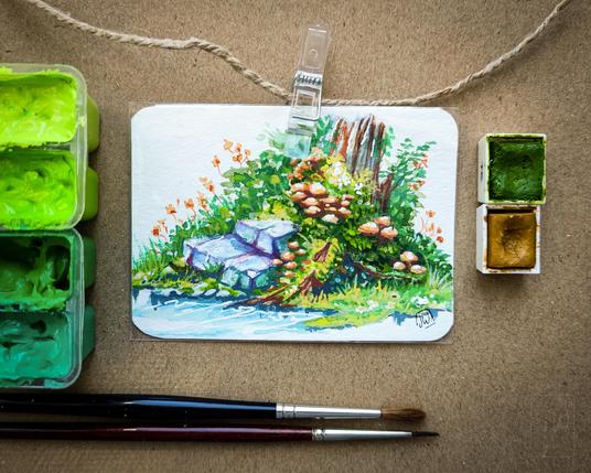 Ein kleines, rechteckiges Aquarellbild mit abgerundeten Ecken ist mit einer transparenten Klammer an einer Juteschnur befestigt. Es zeigt eine lebendige Waldszene mit einem alten, vermoosten Baumstumpf, vielen orangefarbenen Pilzen, saftigem grünen Gras, wilden Blumen und ein paar violett-grauen Steinen am Rand einer kleinen Wasserfläche. Das Bild ist sehr detailreich und leuchtend koloriert. Links und rechts neben dem Bild sind geöffnete Aquarellnäpfchen zu sehen – in Grüntönen. Unten im Bild liegen zwei Pinsel auf dem braunen Untergrund.