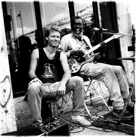 A younger white man wearing a tank talk and jeans sits next to an older white man with an electric guitar and wide open mouth with a microphone in front and some sound gear with a window with reflections in back.