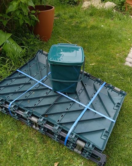 A green flat pack garden composter, bound in blue tape, lying on a lawn. A smaller kitchen scrap bin sits on top.