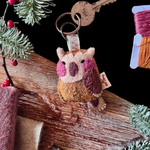 A felt owl keychain photographed on a black and wooden background with decorative plants and felt around. The keychain features an owl with a brown body and beige face and little feathery horns. Its beak and wings are brown, and the tip of its wings are beige. The owl has a beige rectangular tail with a brown little triangle at the base. The owl is looking over its shoulder, its embroidered eyes in a happy squint and its big round cheeks are pink. Attached to its head is an ivory ribbon with brown checkers, linking the owl to a small bronze key ring.