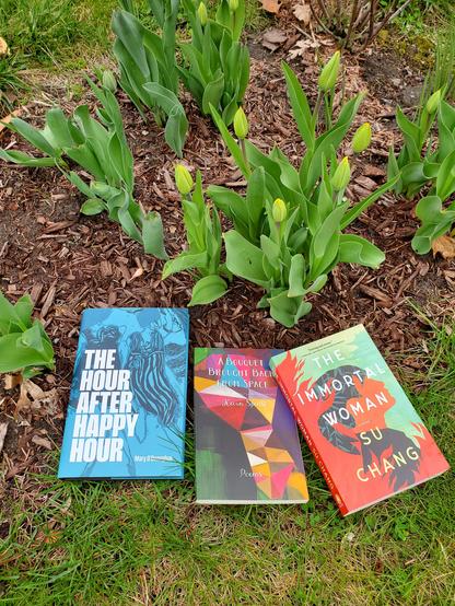 Beautiful books are laid out on the green grass in front of spring tulips getting ready to blossom. The books are: The Hour After Happy Hour by Mary O'Donoghue (Stinging Fly), A Bouquet Brought Back From Space by Kevin Spenst (Anvil Press), and The Immortal Woman by Su Chang (House of Anansi Press)
