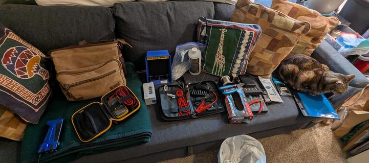 A photograph of a sofa, currently occupied by numerous pillows, a laptop bag, an array of tools and supplies for repairing electronics, and one cat.