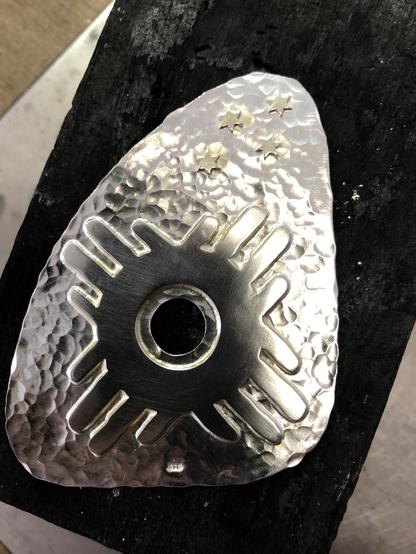 A metallic, hammered piece with a triangular shape features a central hole and decorative designs, including rays and four small stars. It rests on a charcoal surface, highlighting its texture and sheen.