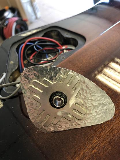 A close-up of a textured metal plate with a central hole, resting on a wooden guitar surface. Beneath the plate, wires in red, black, and blue are visible, indicating an electronic component installation. The background features a partially visible tool or object.