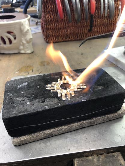 A piece of jewelry is being soldered on a charcoal with flames emerging from the area. Tools and a woven basket are visible in the background.