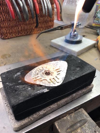 A workbench with a flame torch is being used to heat a metallic piece shaped like a triangle, resting on a black charcoal block. Nearby tools and a glass container are visible in the background.