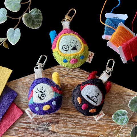 Three felt bag charms representing tamagotchi-like digital toys. One is pink and green with a blob creature standing next to a poop, another one is black and red with a tombstone embroidered on the screen, and a purple, green and red one with an hatching egg on the screen.
