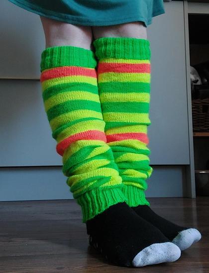 My legs, wearing a pair of legwarmers I knitted. They are bright green, yellow, and orange striped and go up to my knees. There are grey drawers and cupboards behind me, and I also have black socks with grey toes on.