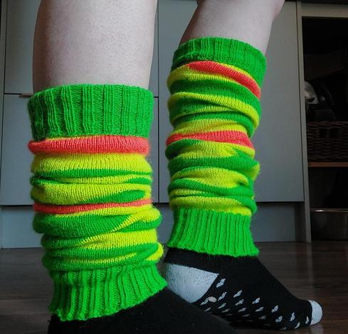 My legs, wearing a pair of legwarmers I knitted. They are bright green, yellow, and orange striped and are loosely scrunched up round my ankles. There are grey drawers and cupboards behind me and I also have black socks with grey toes on.