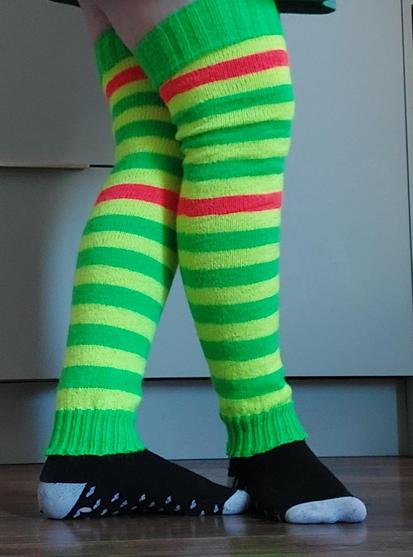 My legs, wearing a pair of legwarmers I knitted. They are bright green, yellow, and orange striped and go up over my knees. There are grey drawers and cupboards behind me and I also have black socks with grey toes on.
