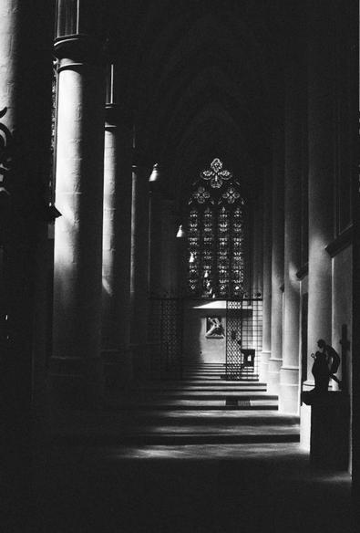 Black and white photo of an aisle in a medieval Gothic church. The columns are half in light and half in shadow and create horizontal shadows across the composition. In the background is an iron gate and a stained glass window. In the foreground is a statue in silhouette.