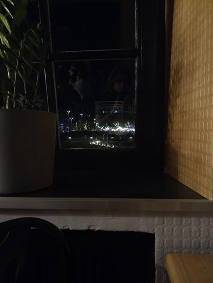 View through a part of a window reveals the other side of a river with a landing, trees and stting lights, some big buildings are barely visible against the night sky.