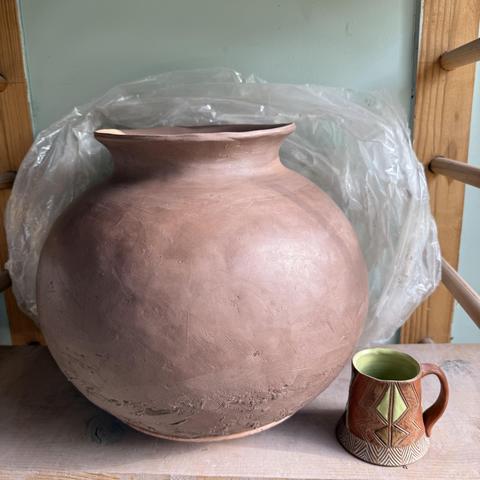 A large, clay pot, unfired and still moist, on a wooden shelf next to a mug, for scale. The pot was coil built with brown clay and is spherical with a wide, short flared neck. The large pot is about 3-4 times the height of the mug and just as wide. The mug is brightly decorated with chartreuse green and earth tones. There is clear plastic bunched behind the large pot, almost framing it. It was pushed back to take the photo. The wall behind is a pale, soft blue-green. 