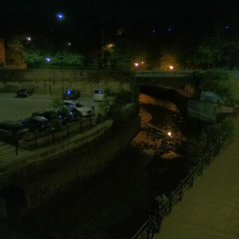 Photograph of the tidal stretch of Ouseburn River at south side of Ouseburn Bridge in Newcastle upon Tyne. To the left (south-west) of the bridge is Cut Bank, and to the right (north-east) is Byker Bank. In the foreground is the pedestrian Riverside Walk and the photo also spans some surrounding built-up areas and trees, with sky in the background. Still image captured from webcam at 03:12 hrs on Saturday 3 May 2025.
