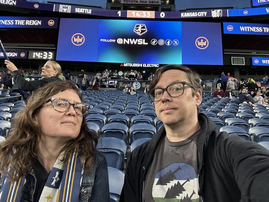 The author and his wife posing in front of a scoreboard reading “Seattle 1 - Kansas City 0”