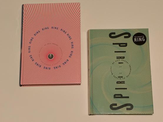 Two books:
- Ring by Koji Suzuki, with cover art showing parallel red and white lines looking down a cylinder with a skull at the bottom
- Spiral by Koji Suzuki with yellow and green spiral cover art
