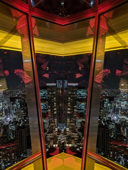 The camera points outside through a window in the Tokyo Tower with the borders being yellow and red. The windows reflect many shapes of objects behind the camera. It's placed right next to a mirror, so the left and right side of the photo look like they're mirrored, cut in the middle. Outside, part of the Tokyo skyline can be seen, illuminated by countless lights. 