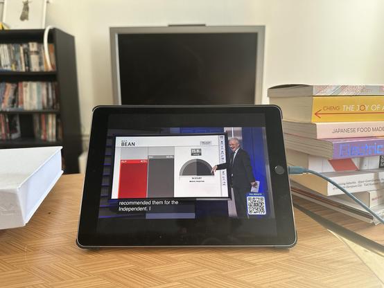 an iPad on a coffee table, its screen showing the ABC election broadcast, with a graph showing a swing from the red party to the grey party. Various books are stacked nearby