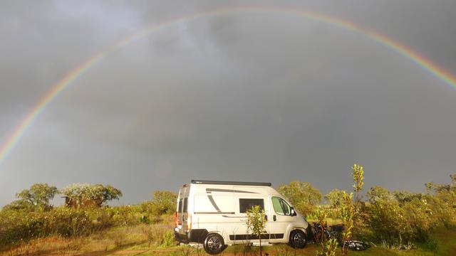 A white camper van at Bubulcus & Bolotas, surrounded by low shrubs and small trees, under a dramatic dark sky. A vivid, full-arc rainbow stretches across the sky, creating a striking contrast against the stormy clouds. A bicycle and some gear are placed near the van, suggesting an off-grid camping adventure in nature.