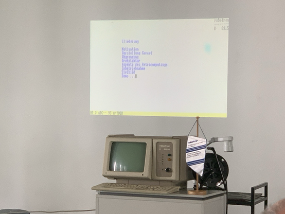 Ein vintage Computer „robotron K 8915“ steht auf einem grauen Tisch. Daneben ein roboterähnlicher Diaprojektor und ein kleiner Wimpel mit der Aufschrift „robotron – VEB Büromaschinenwerk Sömmerda“. An der Wand ist eine Projektion zu sehen mit einer Gliederung für einen Vortrag über das Gerät.