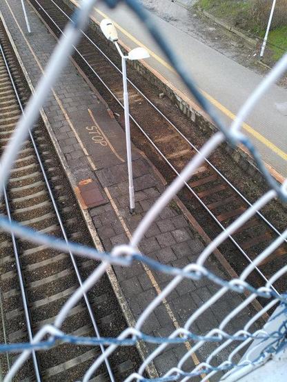 Durch die Maschen eines Zaunes blickt man auf Gleise und einen alten Bahnsteig, auf dem "Stop" geschrieben steht.