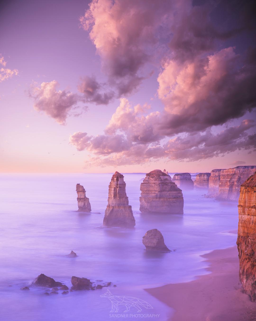 A dreamy photograph of the Twelve Apostles along the Great Ocean Road in Victoria, Australia. Limestone stacks emerge from the misty ocean, with smooth waves lapping at the sandy shore, framed by dramatic cliffs under a pastel sky with soft clouds. A watermark reading "Sandner Art & Photography" is visible in the bottom left corner.
A dreamy photograph of the Twelve Apostles along the Great Ocean Road in Victoria, Australia. Limestone stacks emerge from the misty ocean, with smooth waves lapping at the sandy shore, framed by dramatic cliffs under a pastel sky with soft clouds. A watermark reading "Sandner Art & Photography" is visible in the bottom left corner.